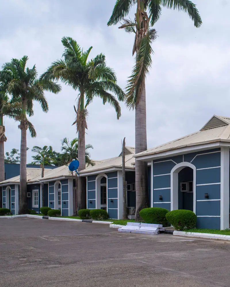 A row of blue single-storey apartments with arched doorways, hedges and palm trees in Abuja, Nigeria.