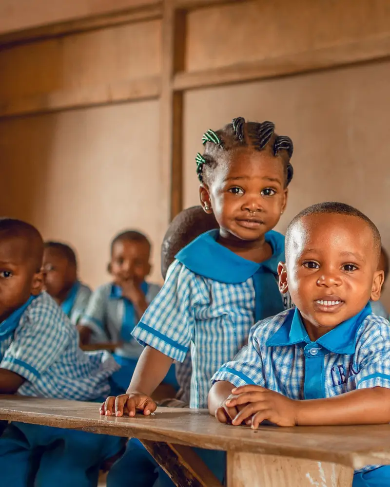 Young pupils in blue and white checked uniforms sitting at wooden desks in a classroom in Nigeria.