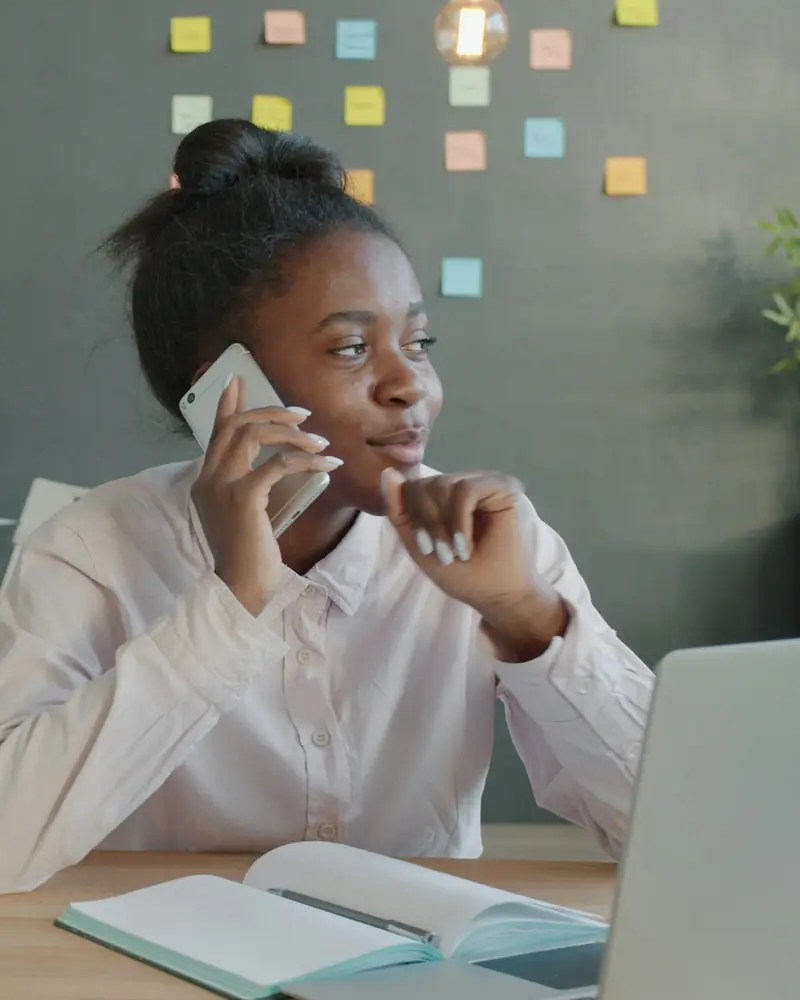 A woman at her office desk, on the phone, with a notebook open in front of her.