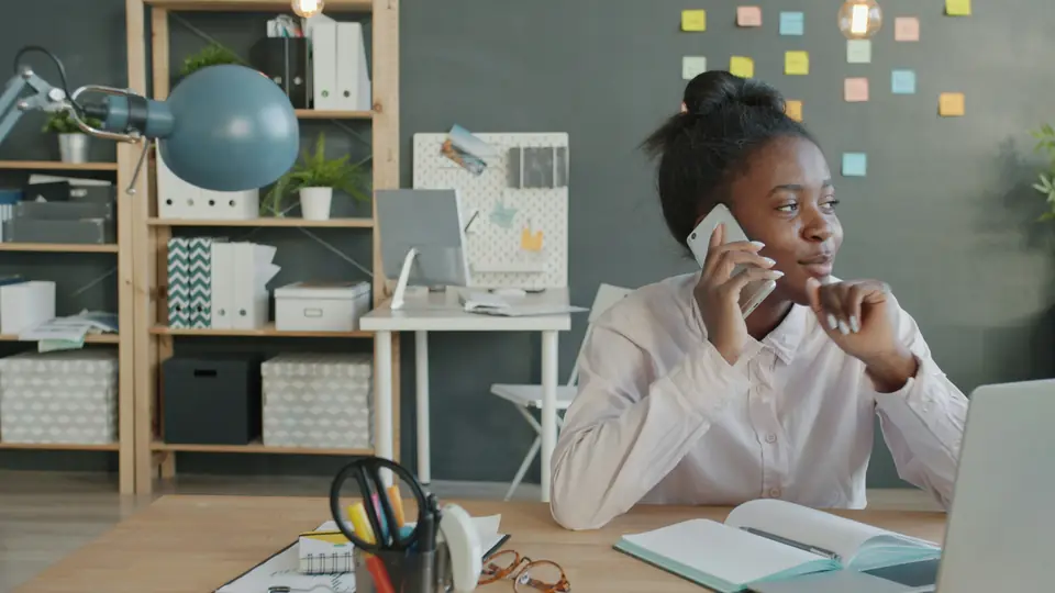 A woman at her desk, smiling a little as she talks on the phone, a notebook open in front of her.