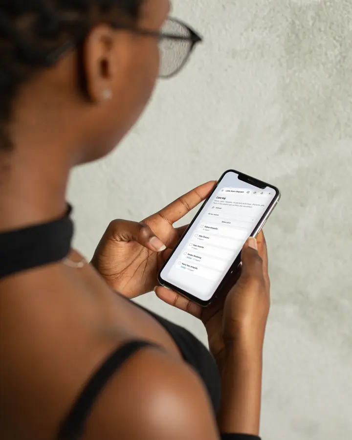 A woman in glasses looking at her phone, which shows a daycare's Care log in Rowsta: the children in her rooms, two of them asleep.