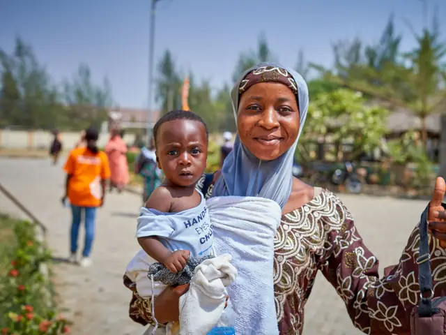 A smiling mother in a headscarf holding her baby outdoors in Keffi, Nigeria.