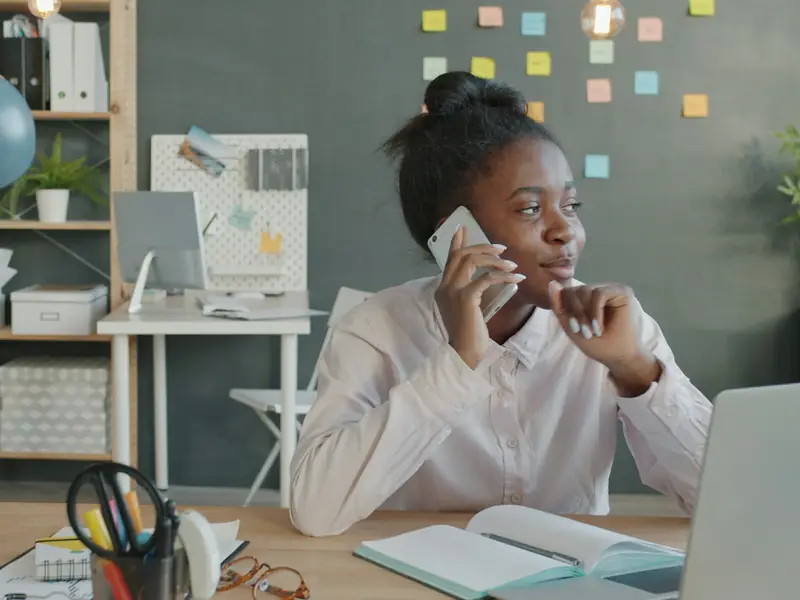 A woman at her office desk, on the phone, with a notebook open in front of her.
