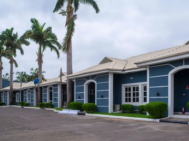 A row of blue single-storey apartments with arched doorways, hedges and palm trees in Abuja, Nigeria.