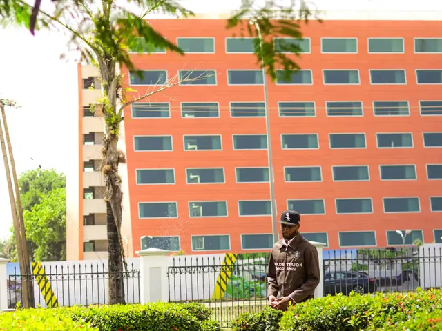 A security officer in uniform walking along a garden path in front of a tall building in Abuja, Nigeria.