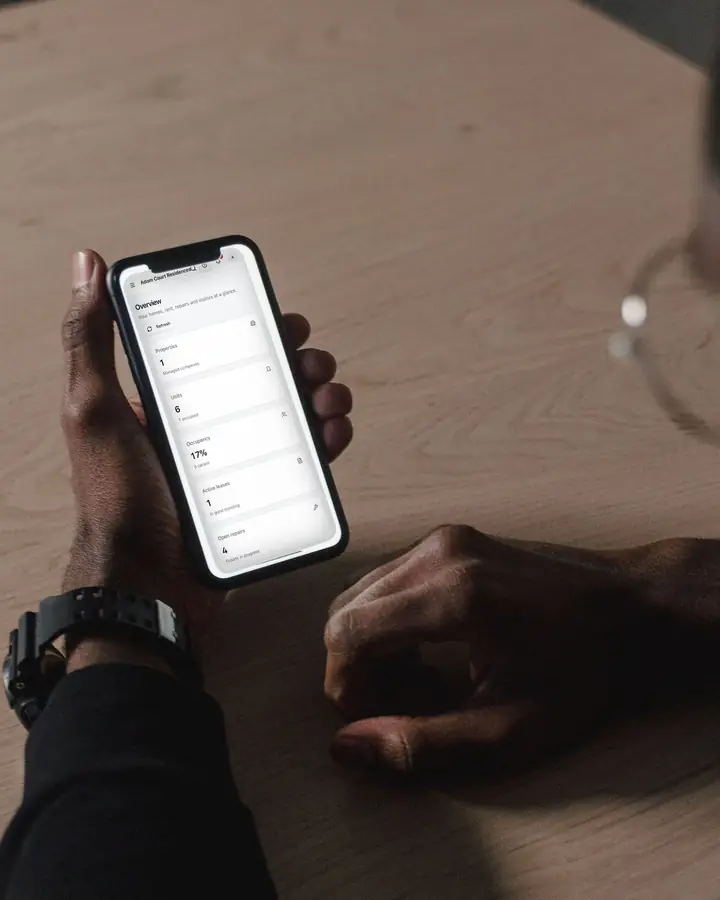 A man at a table holding his phone, which shows an estate's Overview in Rowsta: properties, units, occupancy, leases and open repairs.