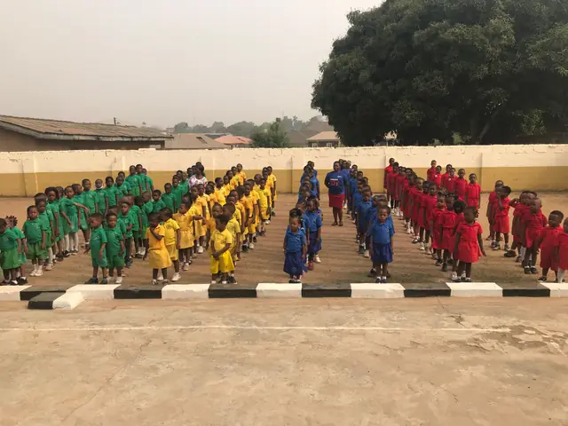 Pupils in green, yellow, blue and red uniforms standing in rows on a school ground for morning assembly in Warri, Nigeria.