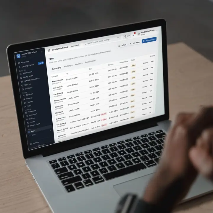 A man at a wooden desk working on a laptop that shows a school's Fees page in Rowsta: what each pupil's family owes, with first-term tuition overdue.