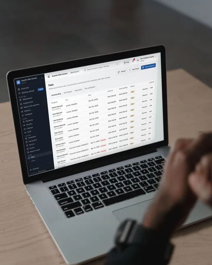 A man at a wooden desk working on a laptop that shows a school's Fees page in Rowsta: what each pupil's family owes, with first-term tuition overdue.