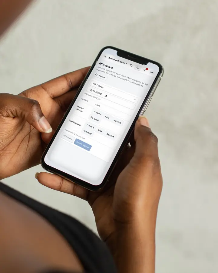 A woman holding her phone in both hands, taking a class register in Rowsta: each pupil with Present, Late, Absent and Excused.